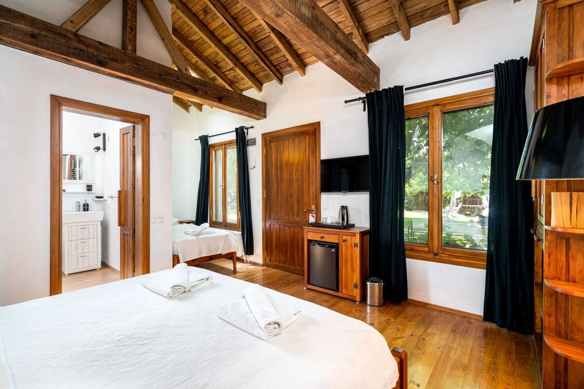 A bedroom with a large bed, wooden beams, a small kitchenette, and a view of greenery outside.