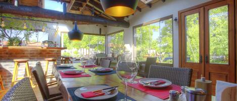 A dining area with a long wooden table set for a meal, surrounded by wicker chairs, and large windows offering a view of greenery outside.