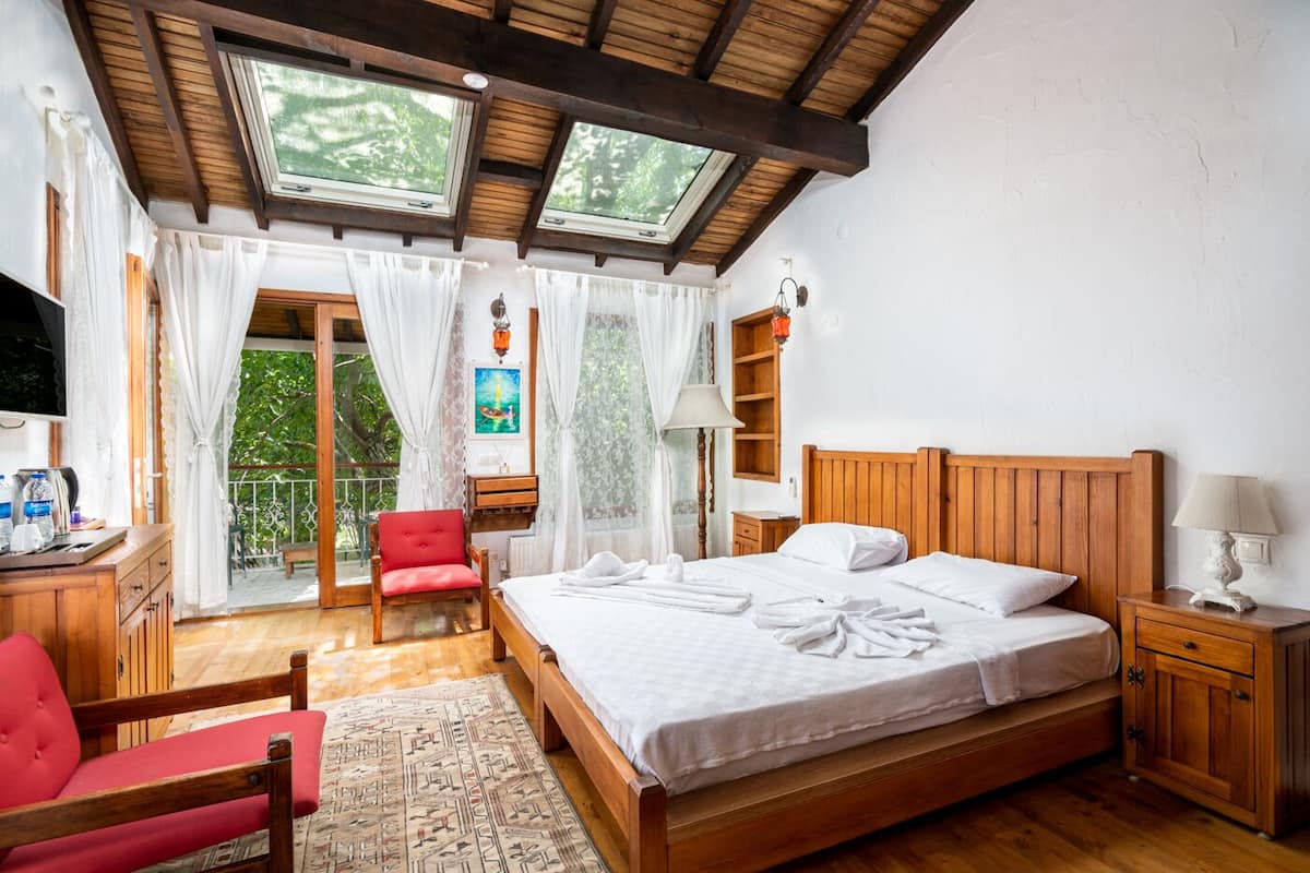 A bedroom with a wooden bed, a red chair, a wooden desk, a TV, and a balcony with a view of greenery.