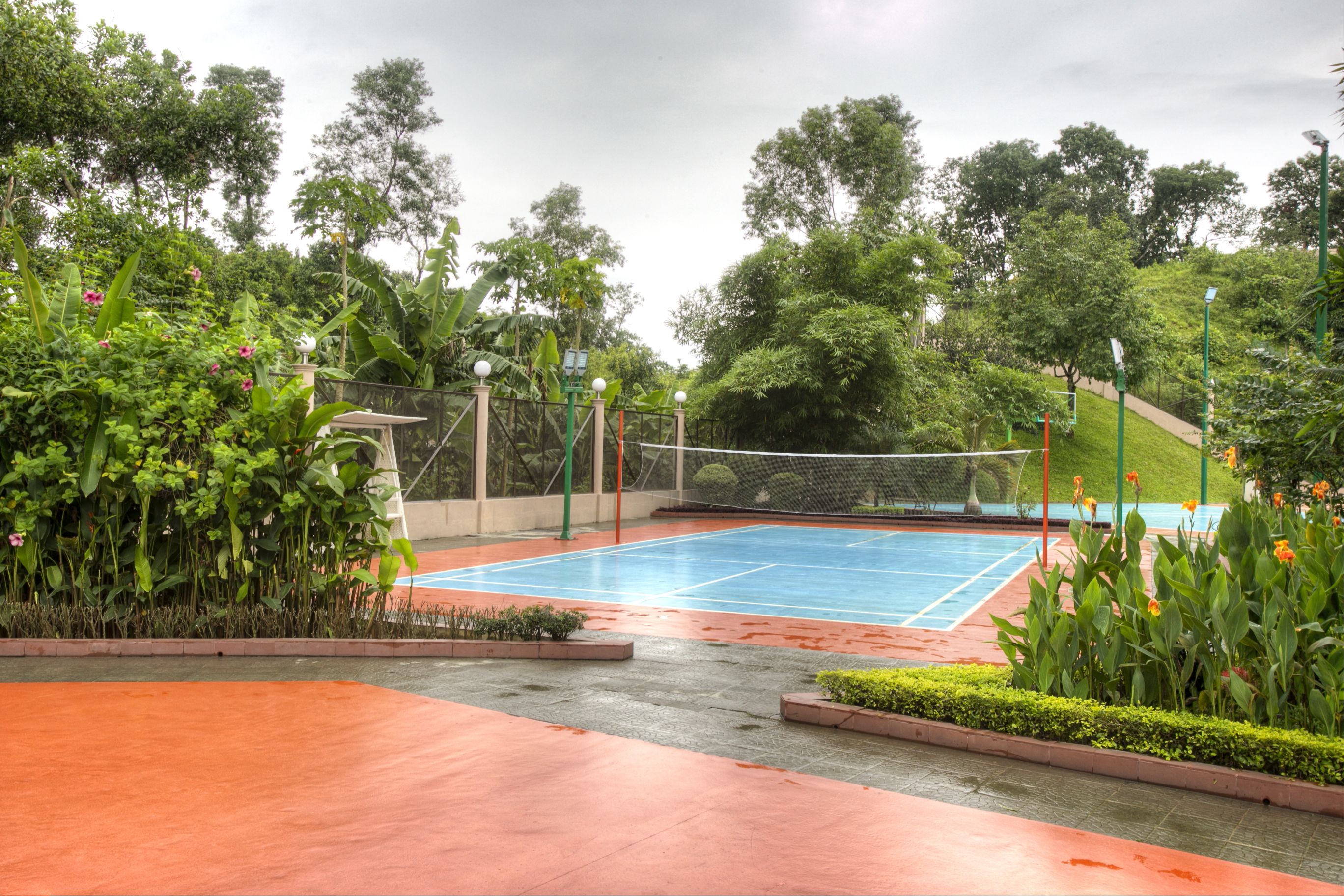 An outdoor tennis court surrounded by lush greenery and a paved walkway.