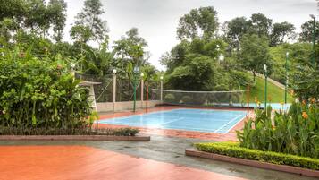 An outdoor tennis court surrounded by lush greenery and a paved walkway.