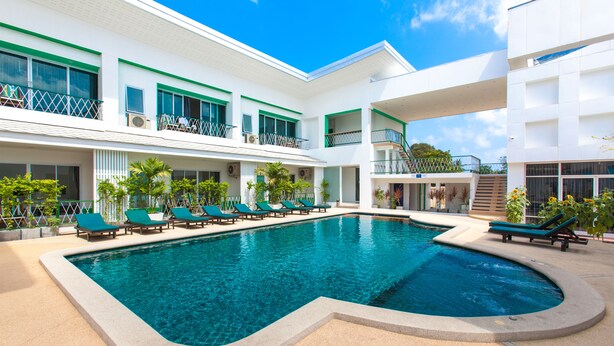 Una piscina con sillones de descanso, un edificio blanco con balcones y un cielo azul despejado.