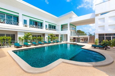 A swimming pool with lounge chairs, a white building with balconies, and a clear blue sky.