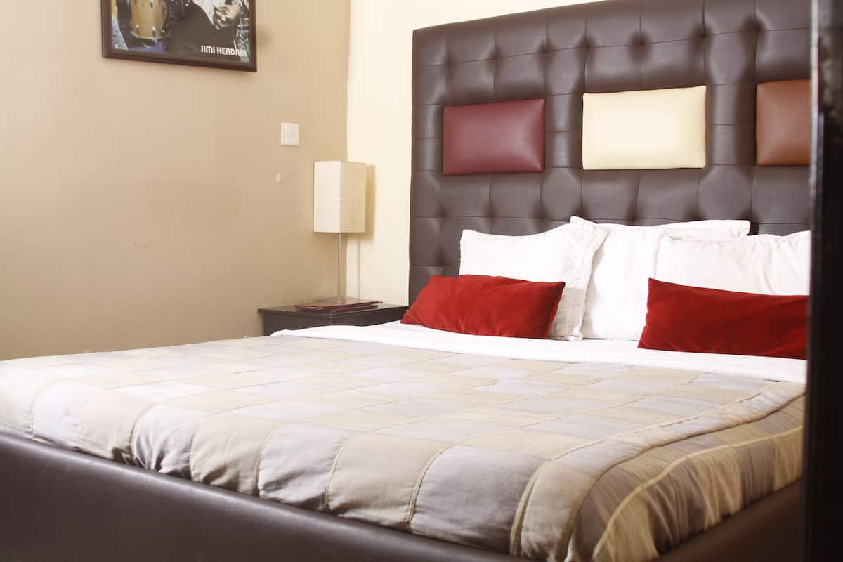 Una habitación de hotel con una cama grande, un cabecero con varios almohadones, una mesita de noche y una lámpara de pared.