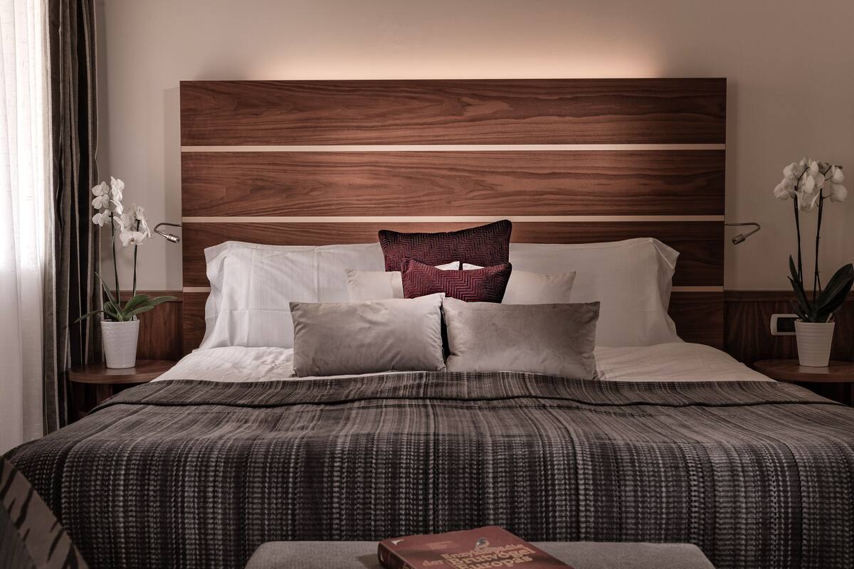 Een net opgemaakt bed met een donkere, patroonrijke spreidekker, een houten hoofdbord en twee witte orchideeën aan weerszijden.
