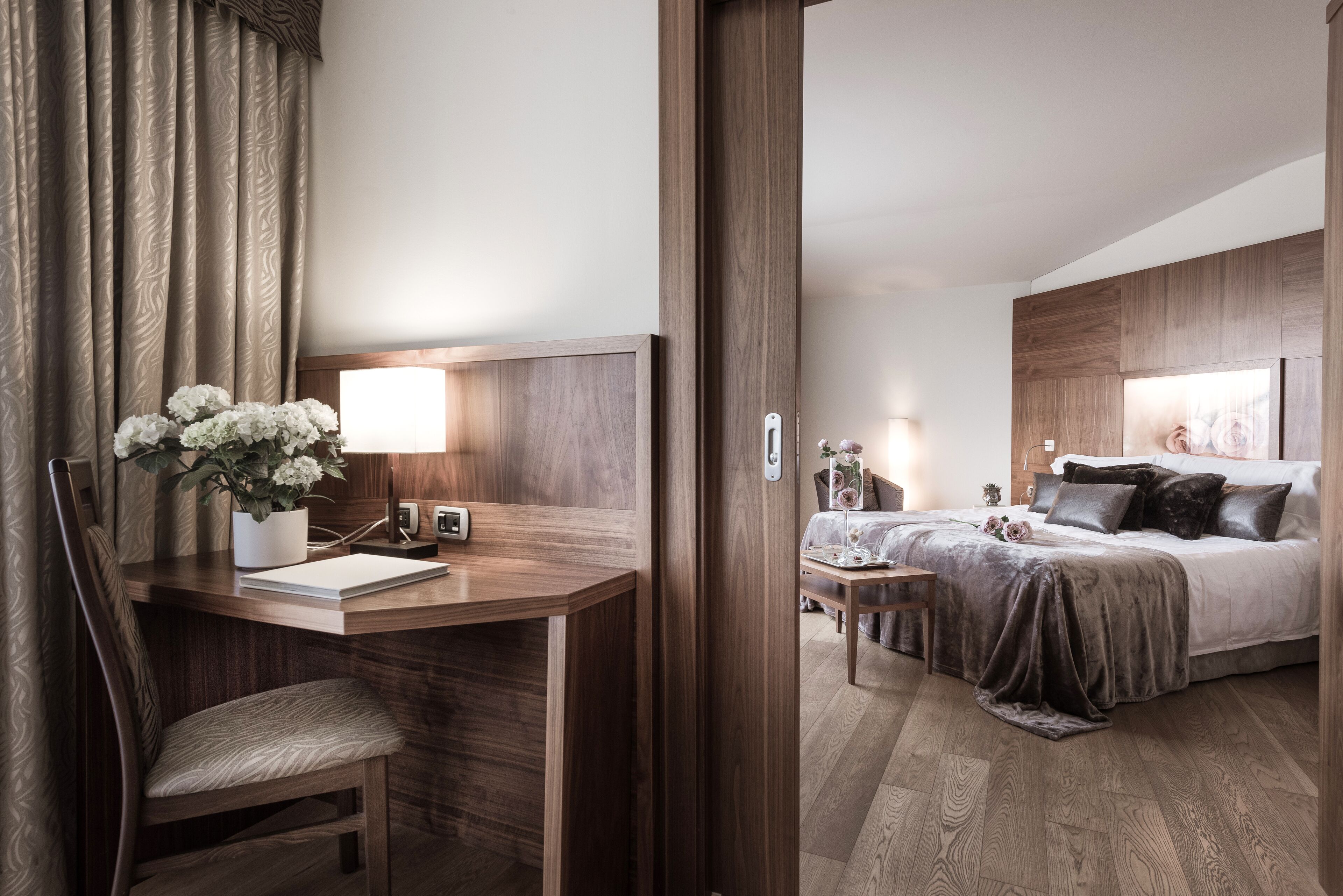 A hotel room with a wooden desk, a chair, a bed with a dark bedspread, and a bedside table with a lamp.