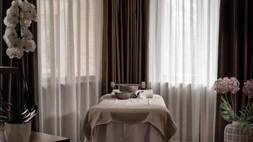 Une chambre avec un lit recouvert d’une couverture beige, une table de chevet avec un bol et un livre, et une plante en pot sur la gauche.