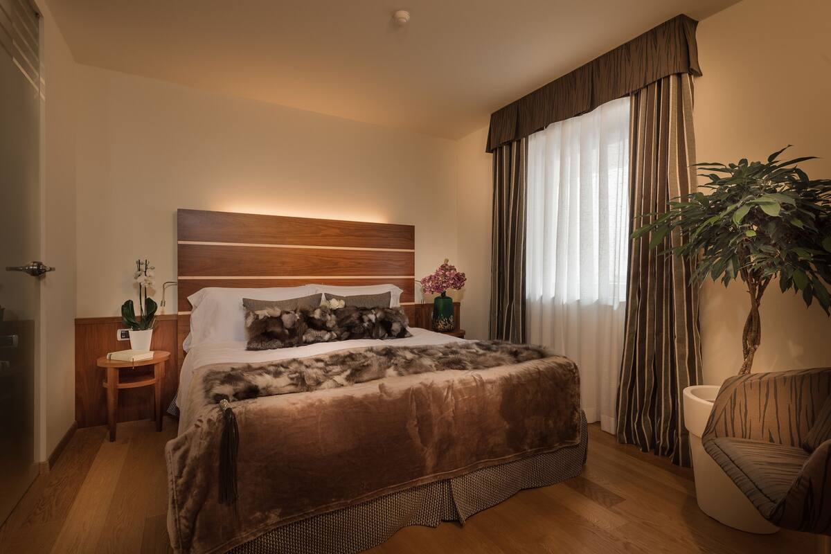 Een slaapkamer met een houten hoofdbord, een bed met een bruine deken, een nachtkastje met een vaas, een stoel en een grote plant.