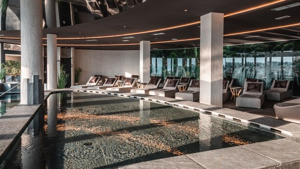 Un espace piscine intérieur moderne, doté de coins salon confortables et offrant une vue sur la ville grâce à de grandes fenêtres.