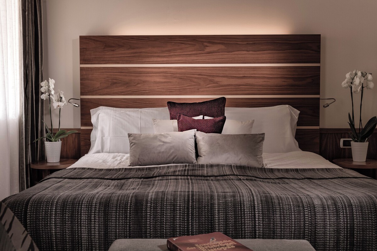 Un lit bien fait, recouvert d’une couvre-lit à motifs, avec une tête de lit en bois et deux tables de chevet ornées de plantes en pot.