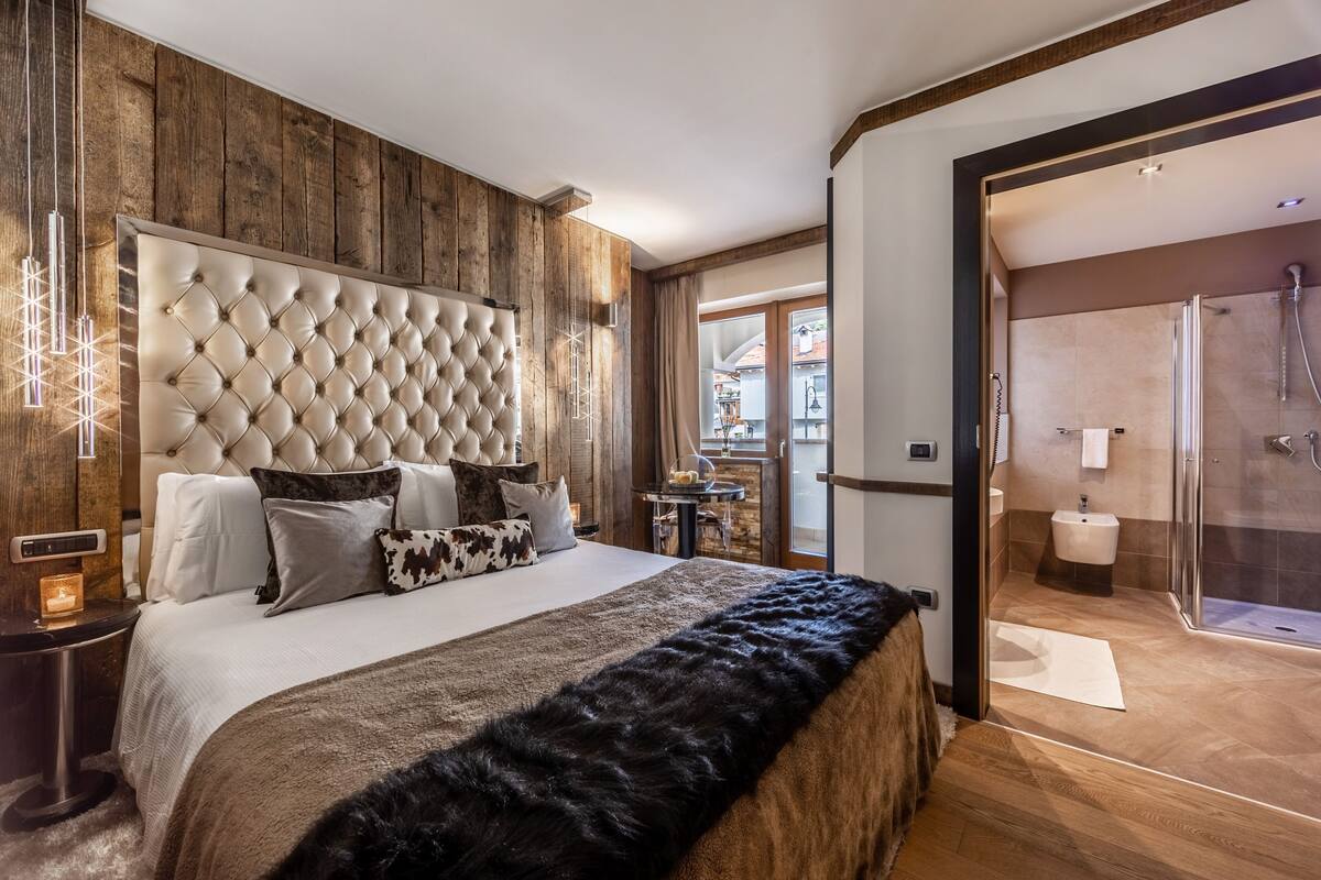 Een slaapkamer met een gestoffeerd hoofdeinde, een groot bed met meerdere kussens, een bont plaid, een nachtkastje met een lamp en uitzicht op de badkamer met een douche.