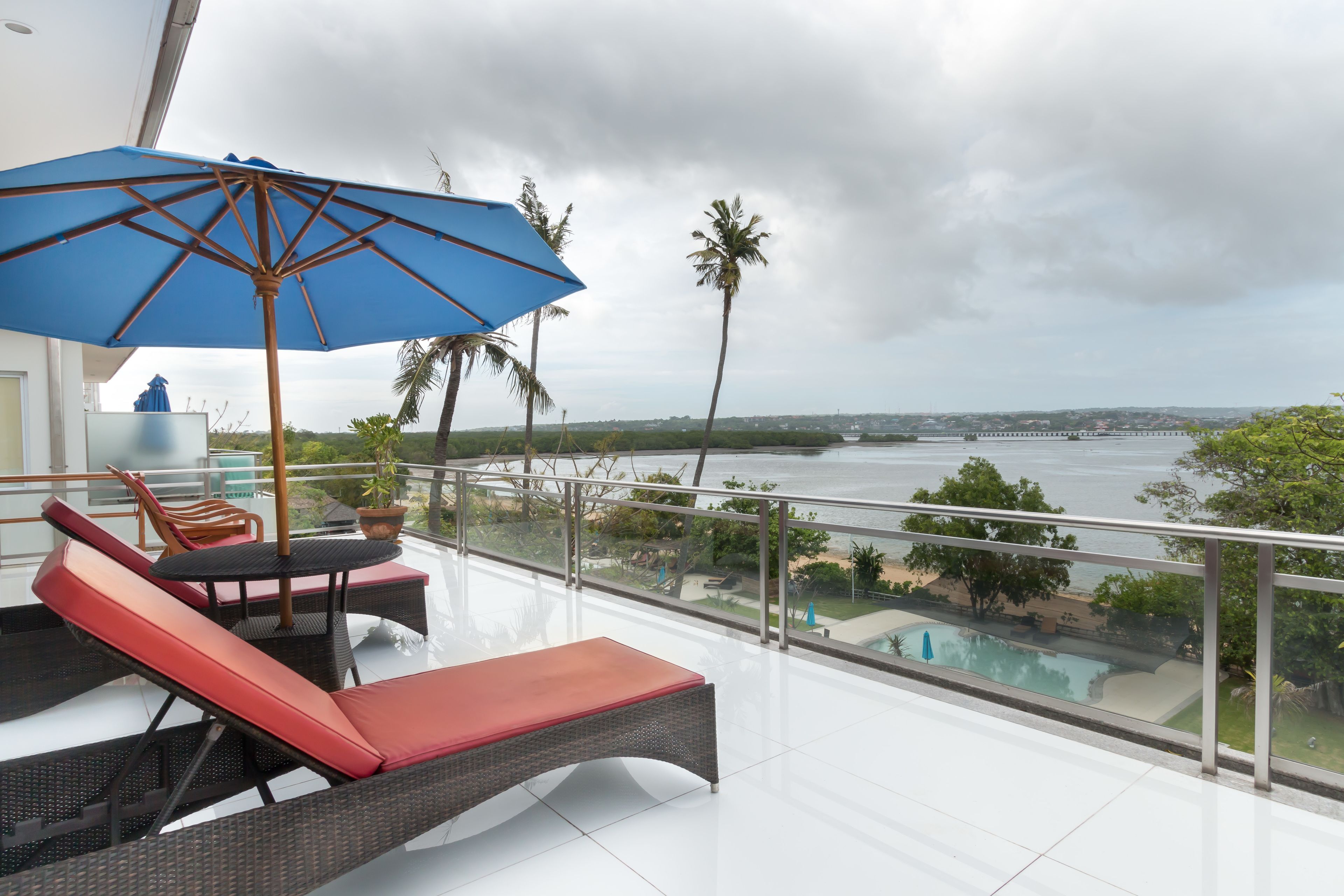 A balcony with wicker furniture and a blue umbrella, overlooking a pool and palm trees.