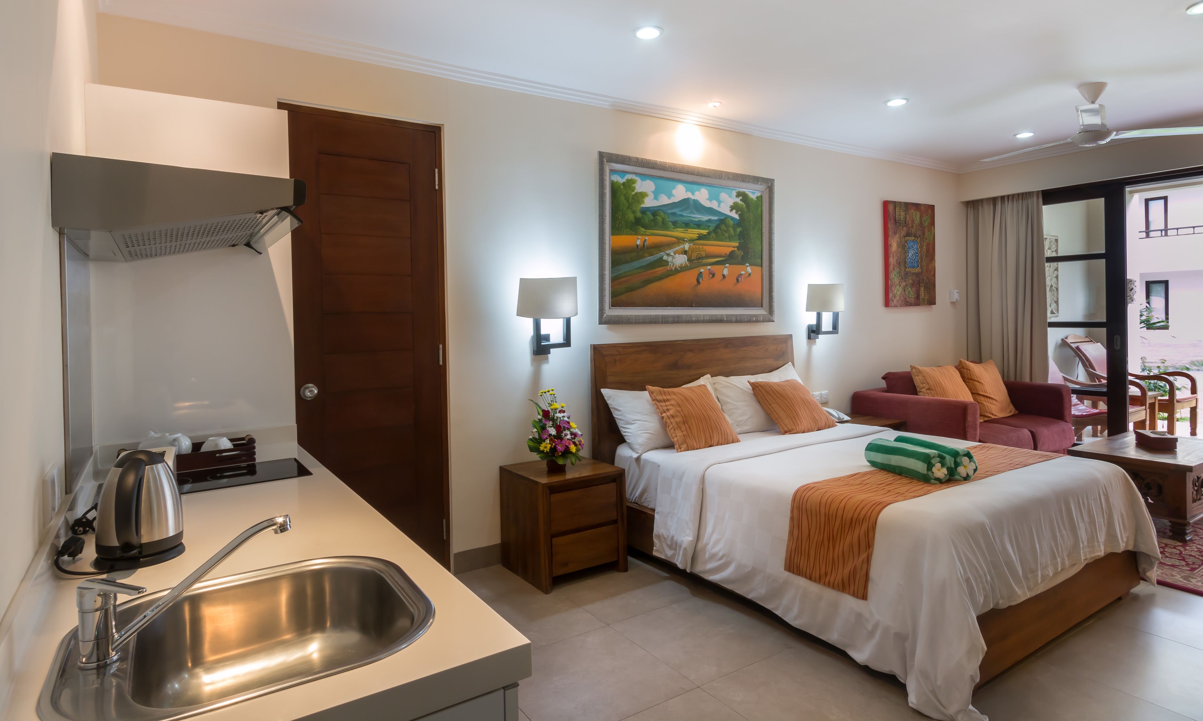 A hotel room with a bed, bedside tables, a desk, a sink, and a painting on the wall.