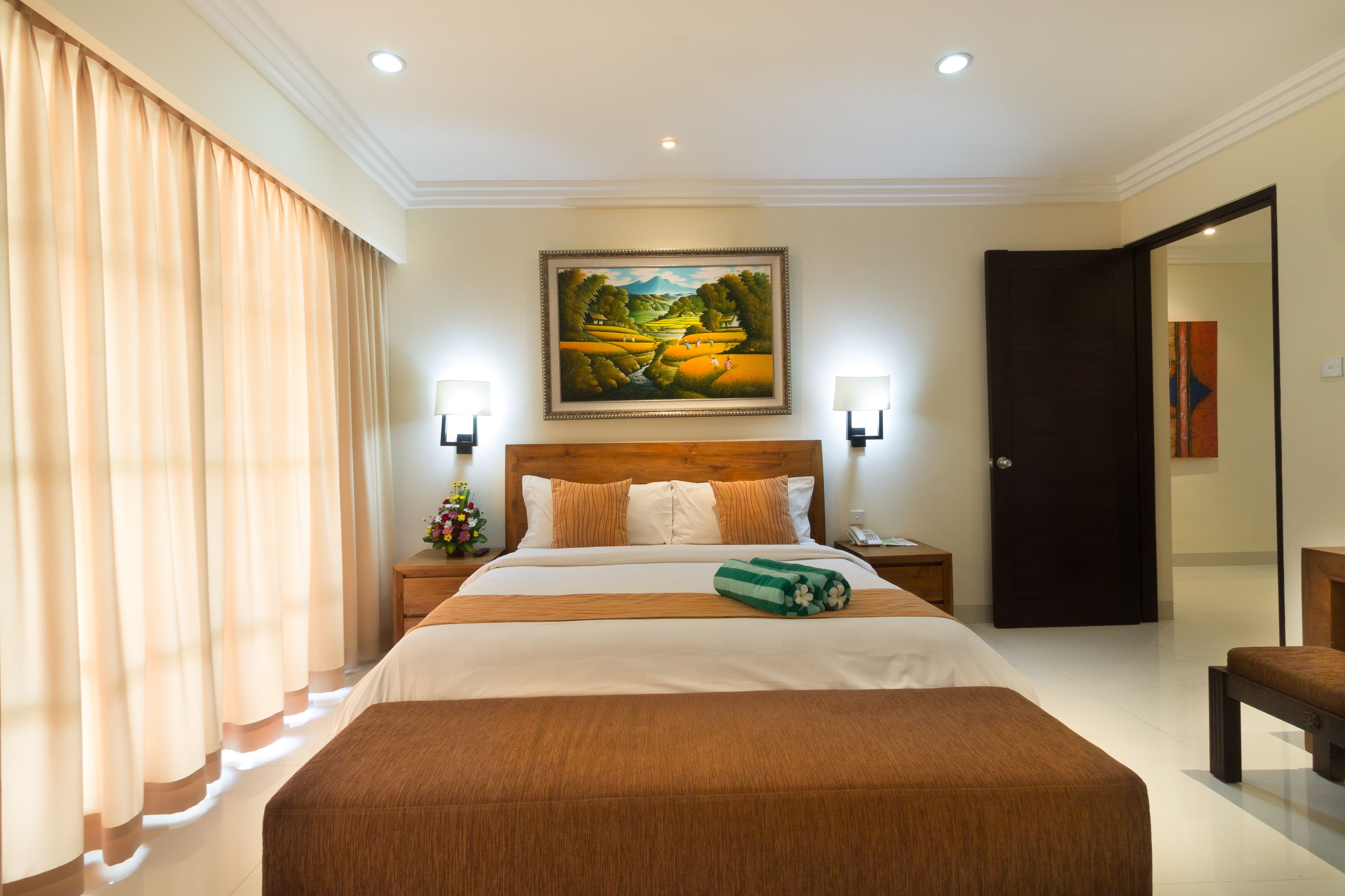 A hotel room with a large bed, a wooden headboard, a painting above the bed, and a nightstand with a lamp.