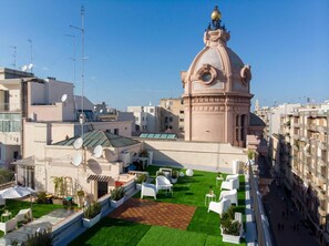 Terraço no telhado com gramado verde, cadeiras de lounge brancas e um edifício com cúpula e torre do relógio.