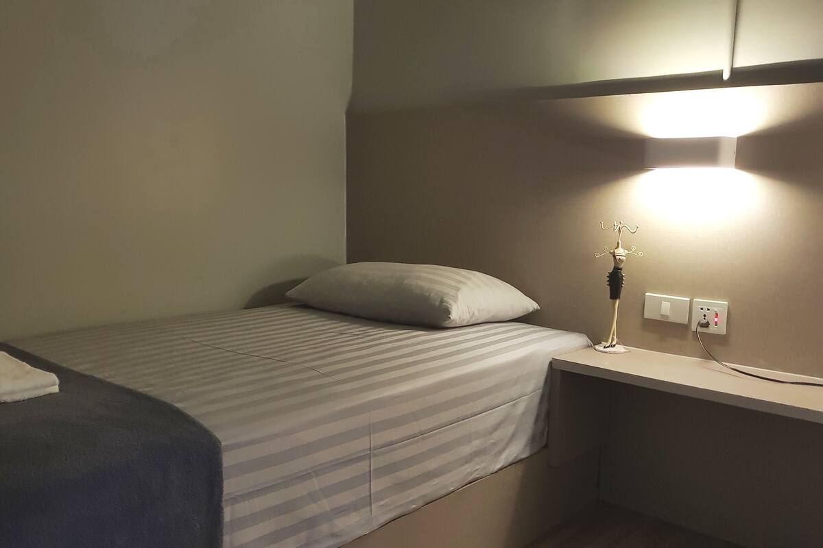 Une petite chambre avec un lit simple, une couvre-lit à rayures, un oreiller blanc, une table de chevet avec une lampe et une prise électrique au mur.