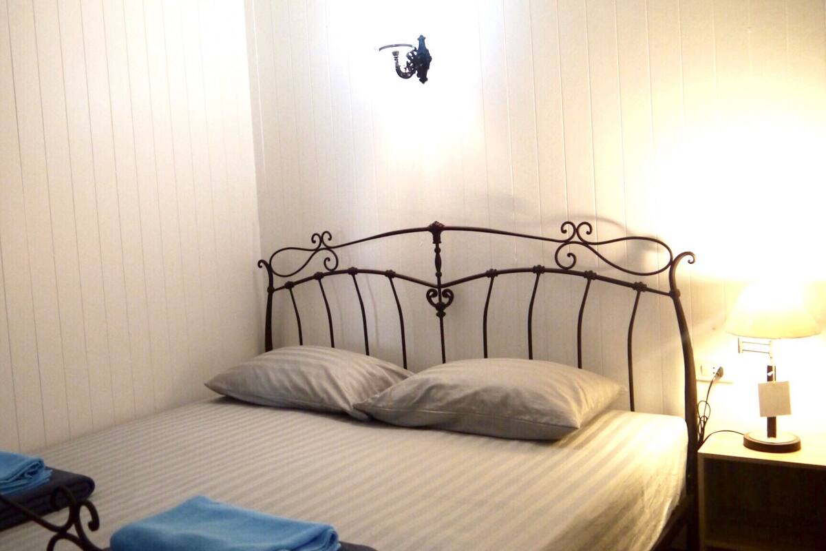 Un lit simple avec une tête de lit en métal, une table de chevet avec une lampe et un luminaire fixé au mur.