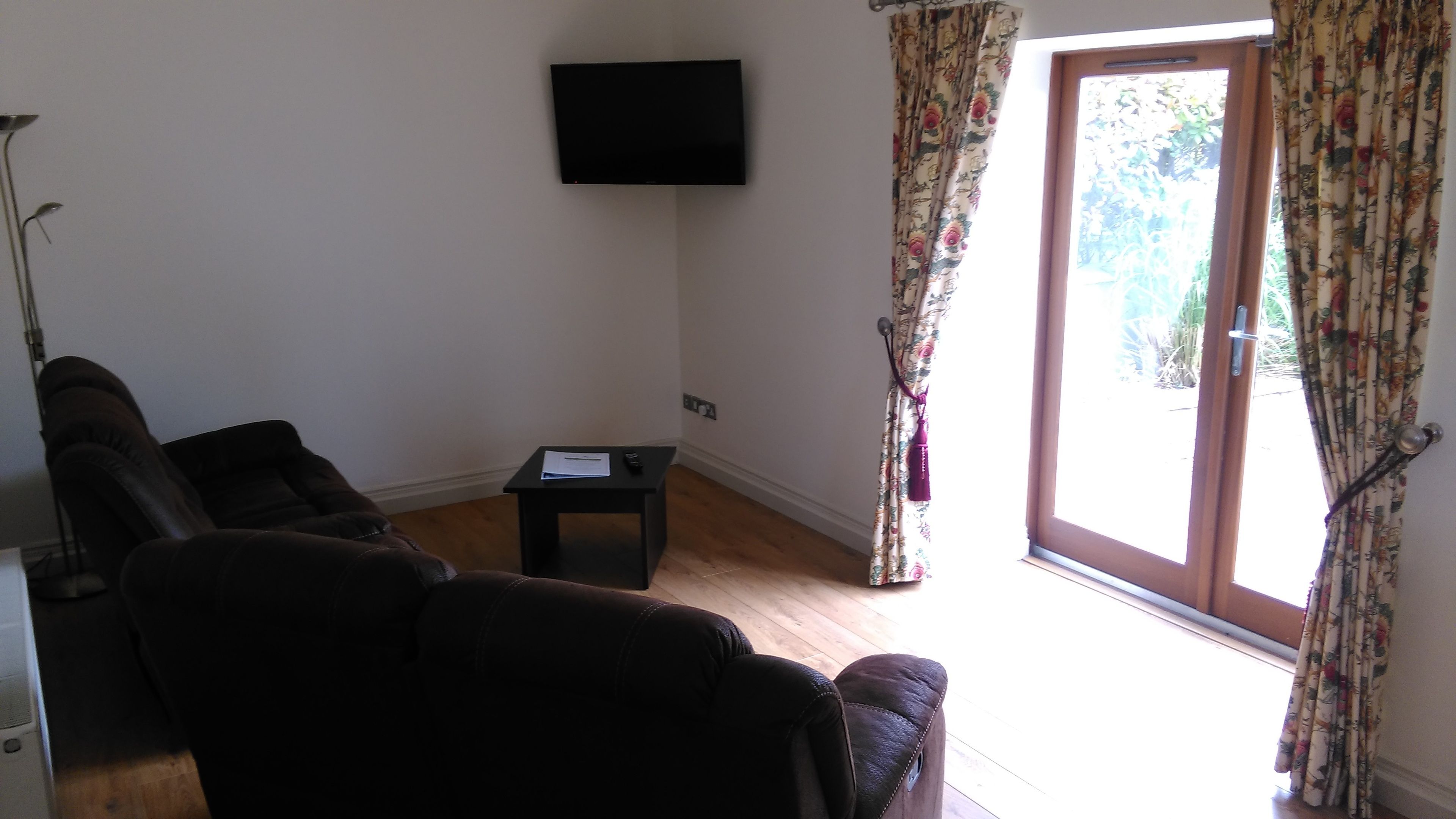 Ein Wohnzimmer mit einer Couch, einem kleinen Tisch, einem an der Wand befestigten Fernseher und einer Tür, die nach draußen führt.