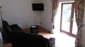 Ein Wohnzimmer mit einer Couch, einem kleinen Tisch, einem an der Wand befestigten Fernseher und einer Tür, die nach draußen führt.