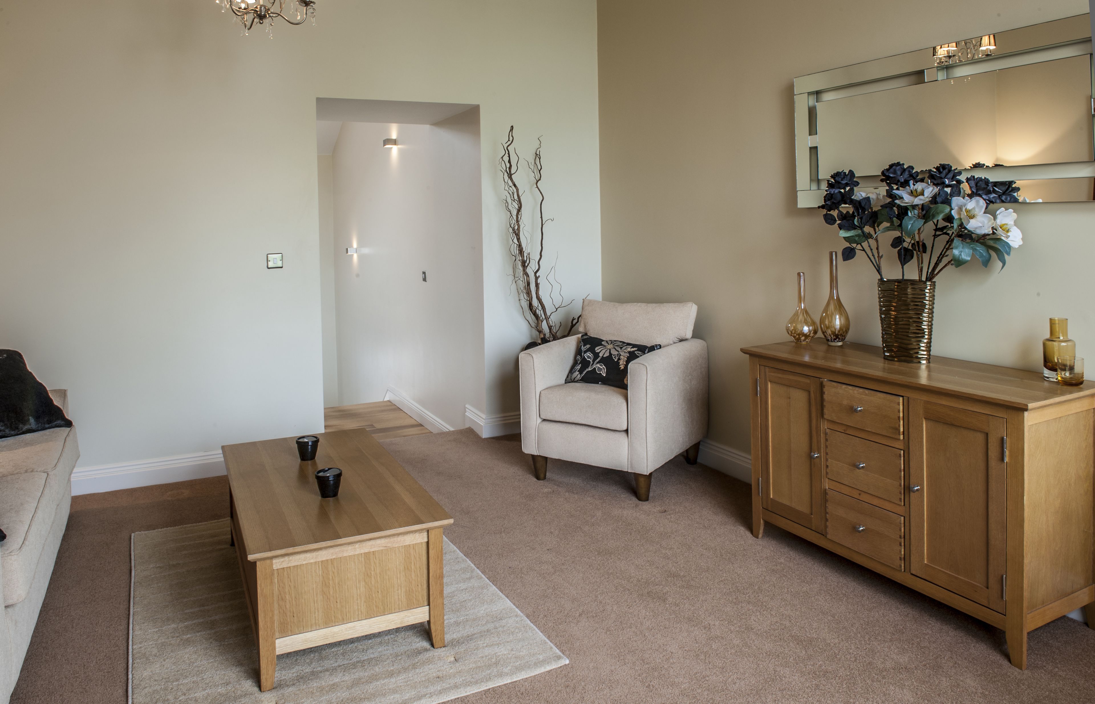 A living room with a beige sofa, a wooden coffee table, a white armchair with a patterned pillow, a wooden cabinet with decorative items, and a mirror above the cabinet.