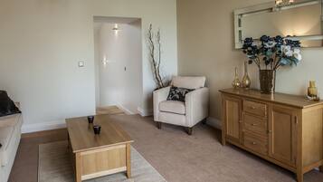 A living room with a beige sofa, a wooden coffee table, a white armchair with a patterned pillow, a wooden cabinet with decorative items, and a mirror above the cabinet.