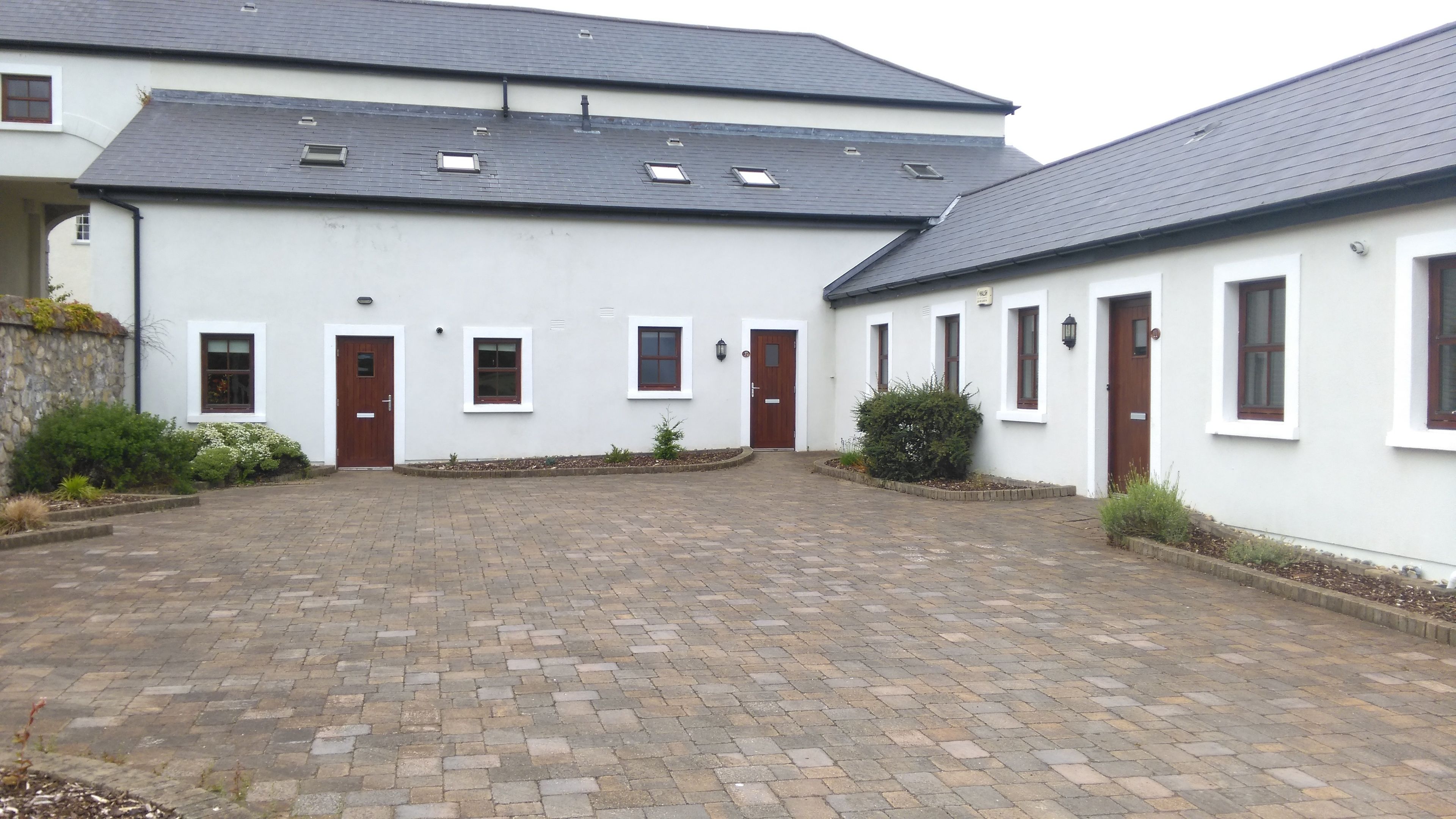 A courtyard with a paved walkway, white buildings with multiple doors and windows, and some greenery.