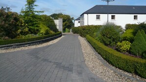 A paved pathway leading to a white building with a dark roof, surrounded by well-maintained hedges and trees. - New Forest Golf Club Apartments (Tyrrellspass)