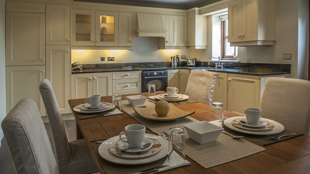 A well-lit kitchen with white cabinetry, a dining table set for four, and a coffee maker visible in the background. - New Forest Golf Club Apartments (Tyrrellspass)