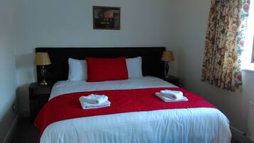 A neatly made bed with a red and white bedspread, two white towels folded on the bed, two bedside lamps, and a floral curtain on the window.