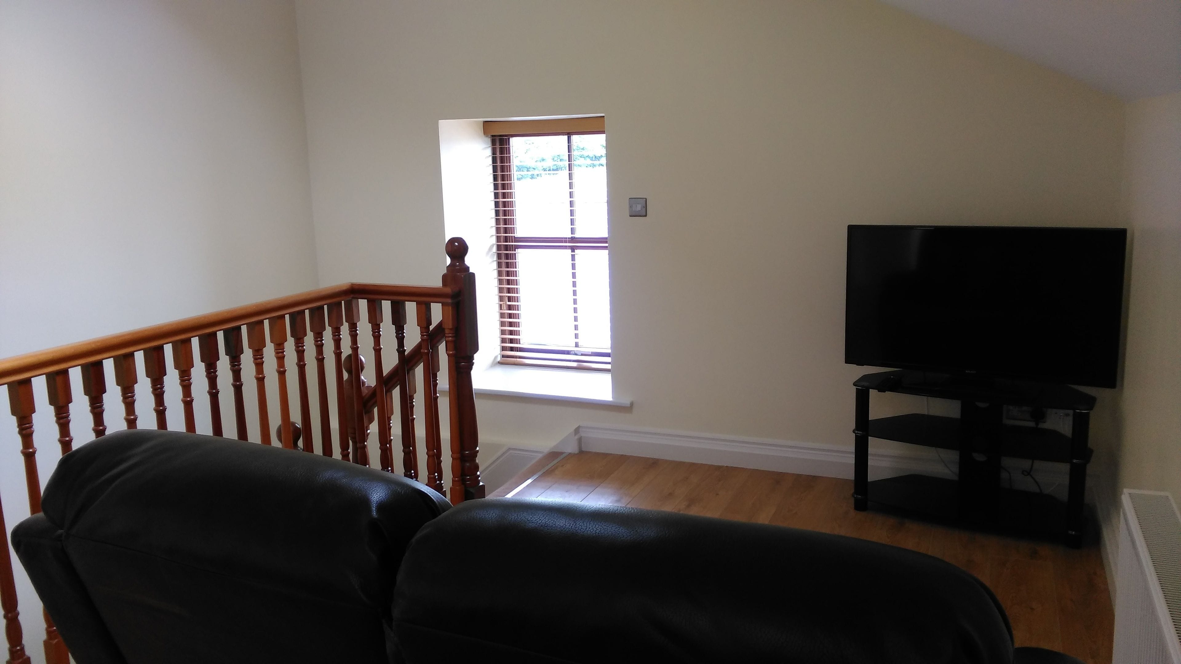 Una habitación con escalera de madera, sofá de cuero negro, televisor sobre un mueble bajo y ventana con persianas.