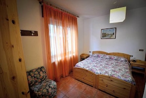 A bedroom with a wooden bed, a chair, a nightstand, and a window with curtains. - Albergo Ristorante Morandi (Valbondione)