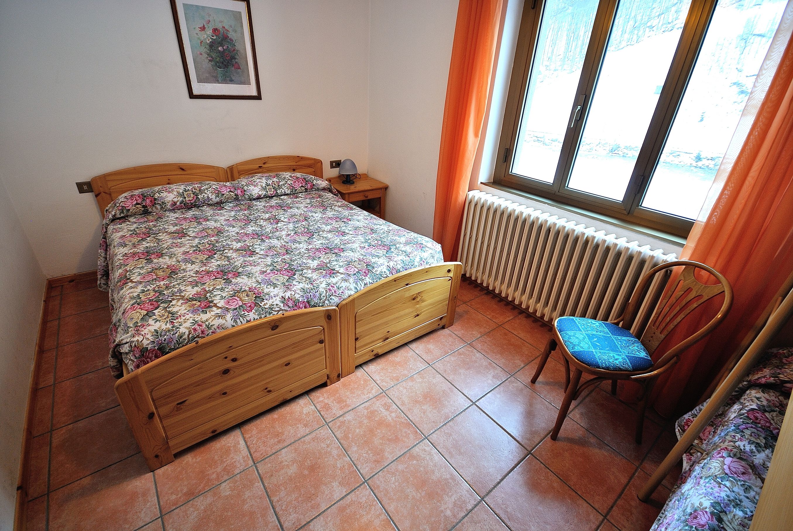 Un dormitorio con una cama doble, una silla, una mesita de noche, un radiador y una ventana con cortinas.