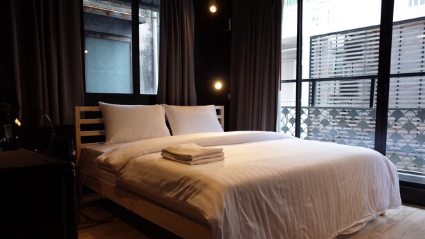 Una cama bien hecha con edredón y almohadas blancos, cabecera de madera y una ventana con vistas a un edificio y un balcón.
