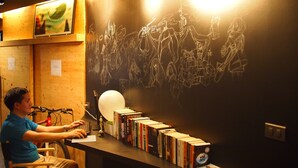 Una persona sentada en un escritorio con una computadora, rodeada de libros, en una habitación con paredes de madera y una pizarra con dibujos.