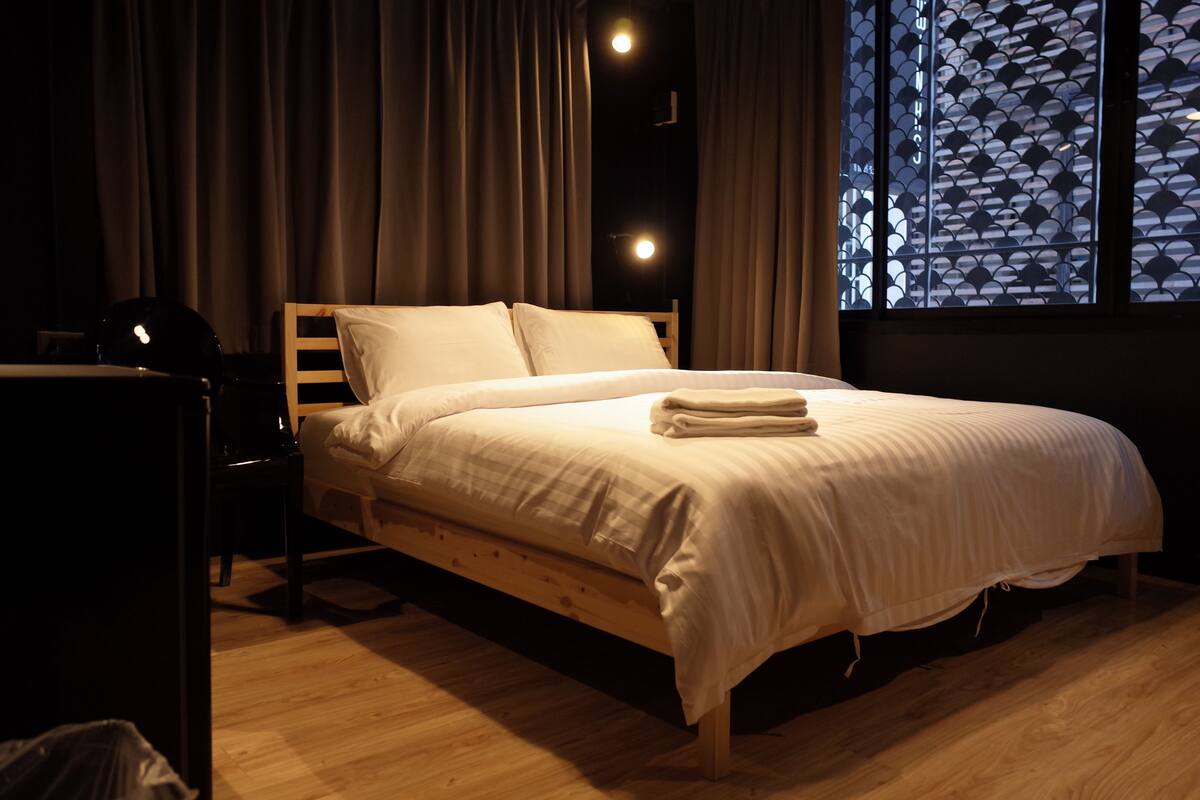 Una cama bien hecha con ropa de cama blanca en una habitación con suelo de madera y una ventana con diseño geométrico.
