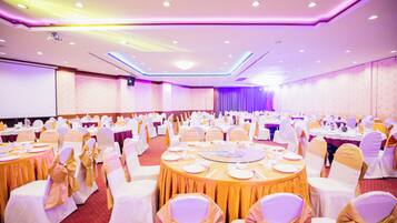 Een feestzaal met ronde tafels gedekt voor een diner, witte stoelen met gouden linten, en een podium aan de voorzijde.