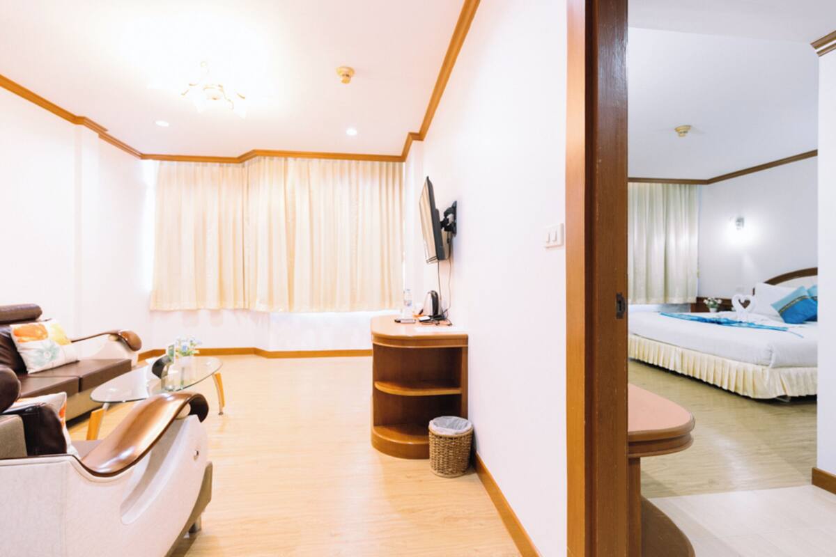 A hotel room with a sofa, a small table, a TV mounted on the wall, a bed with a blue pillow, and a wooden floor.