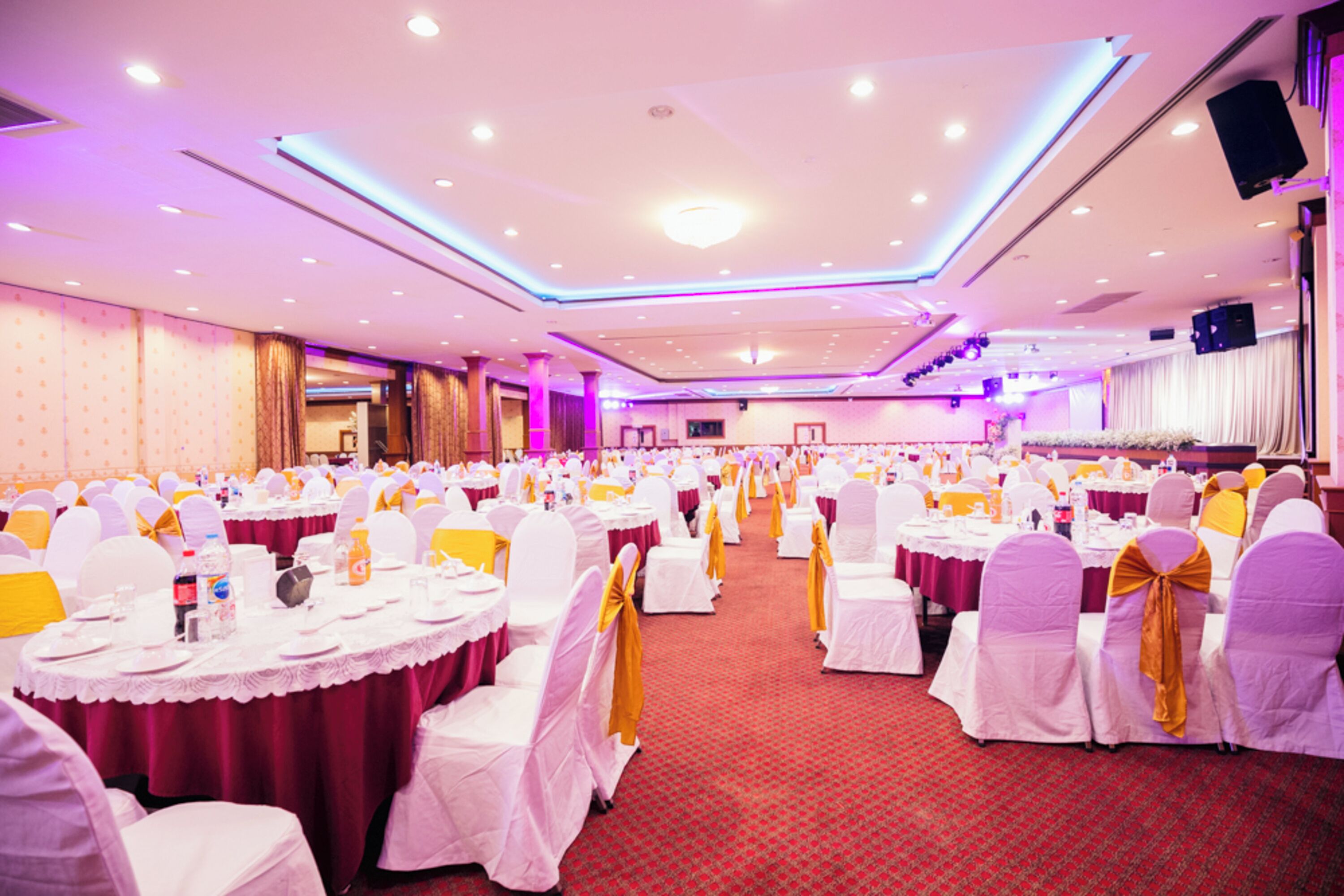 A banquet hall with round tables, white chairs with yellow sashes, and a red carpeted floor.