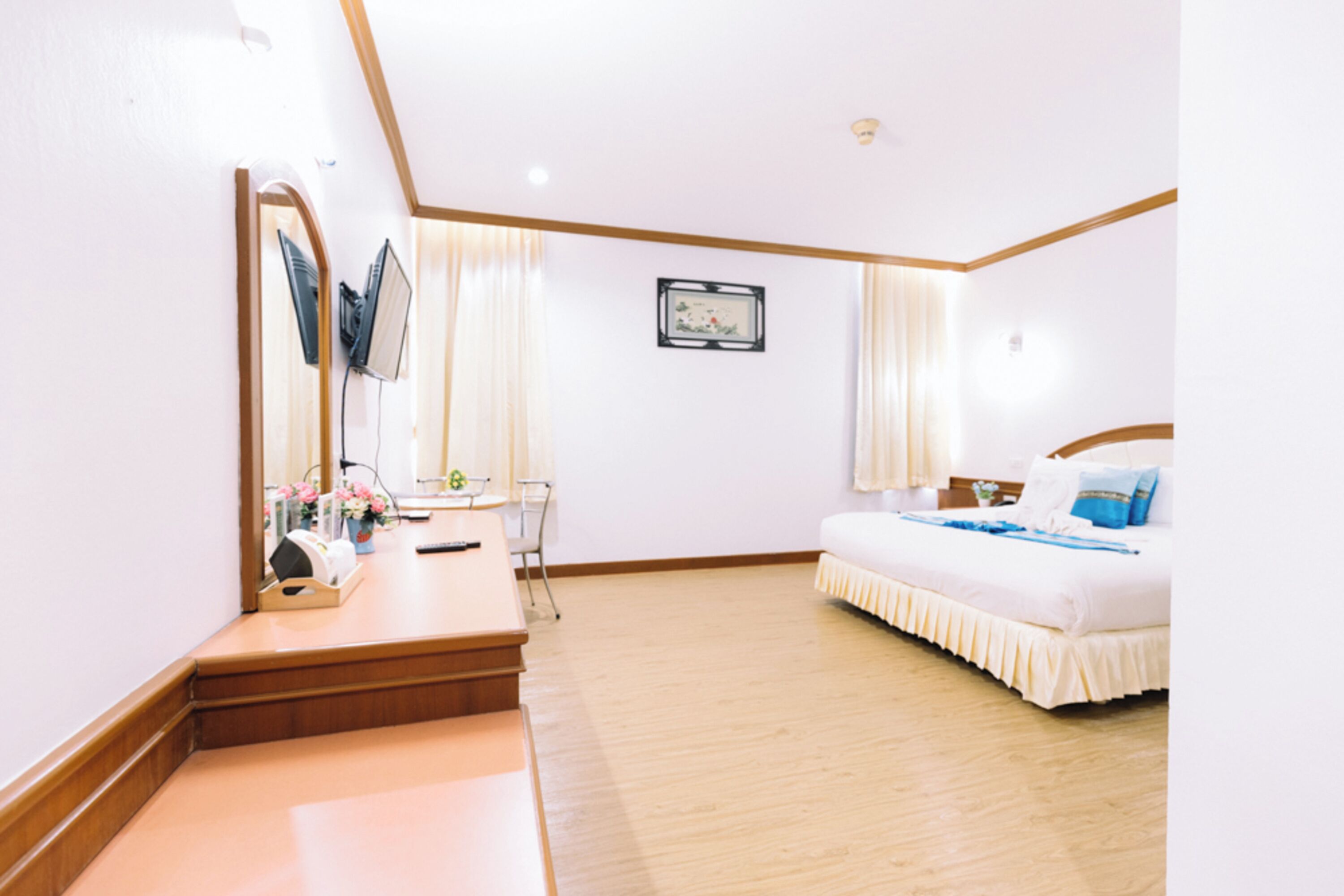 A hotel room with a wooden floor, a bed with white bedding, a desk with a chair, a mirror, and a framed picture on the wall.