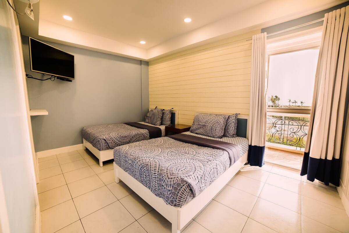Une chambre avec deux lits, un téléviseur fixé au mur et un balcon avec des rideaux.