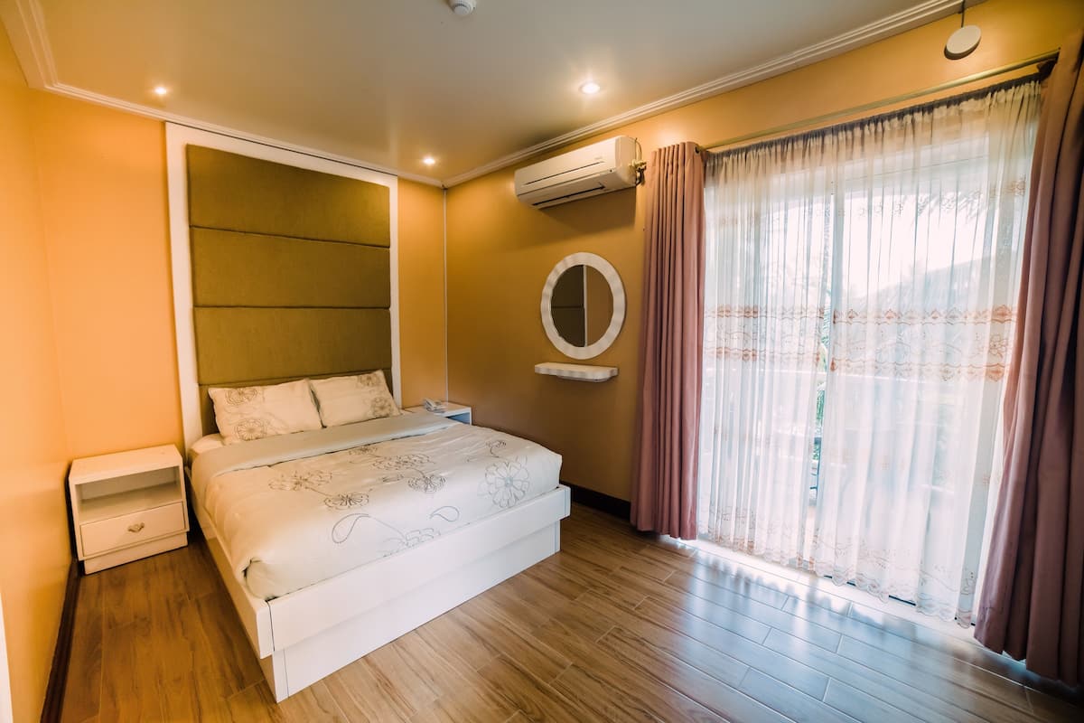 Une chambre à coucher comprenant un lit, une table de chevet, un appareil de climatisation, une fenêtre avec des rideaux et un miroir fixé au mur.
