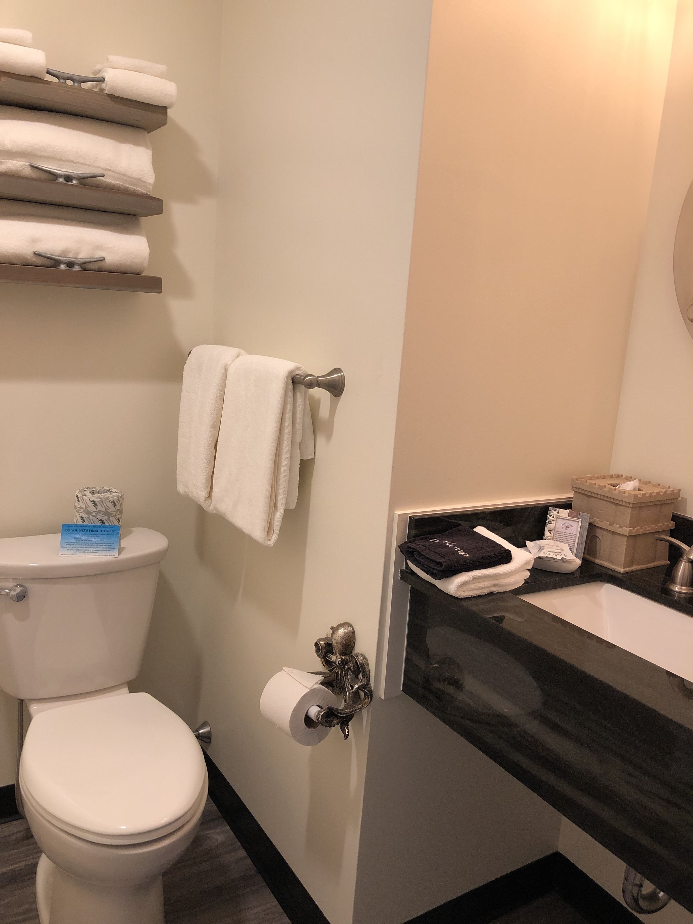 A bathroom with a toilet, a towel rack with folded towels, a paper towel dispenser, and a sink with toiletries.