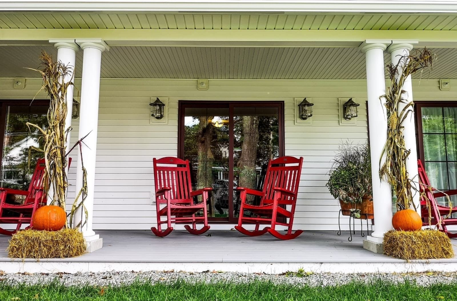 A porch with two red rocking chairs, a white railing, and decorative pumpkins and corn stalks.