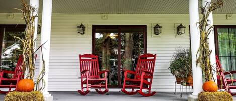 A porch with two red rocking chairs, a white railing, and decorative pumpkins and corn stalks.