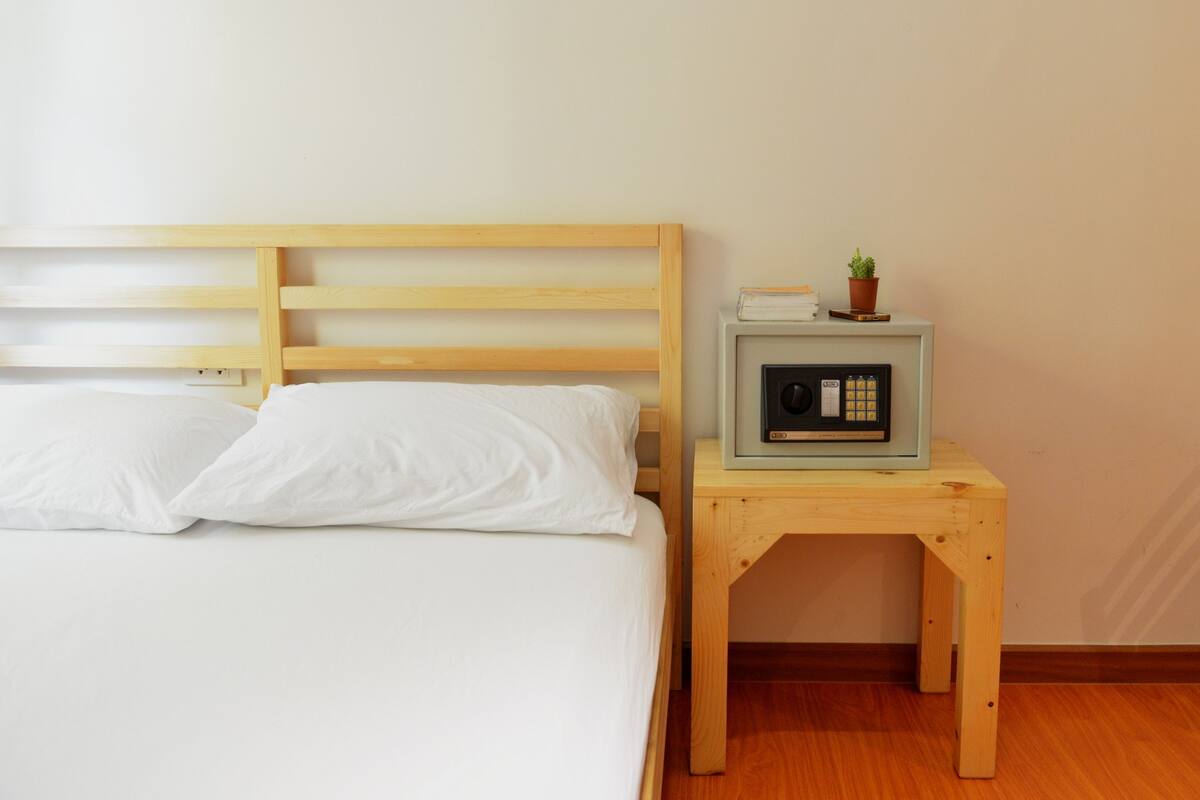 A bed with white linens and a wooden headboard, a small wooden nightstand with a radio, and a potted plant.