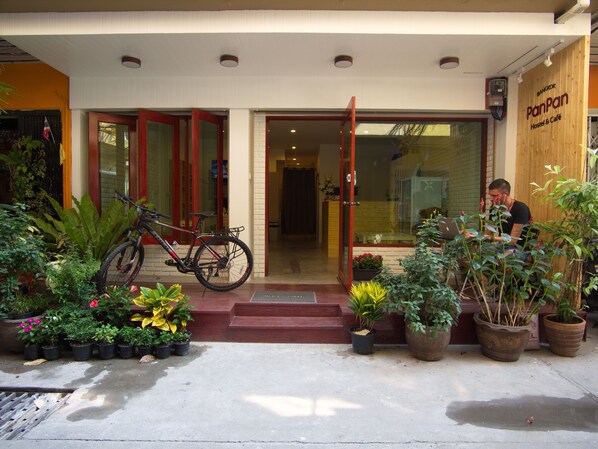 L’entrée d’une auberge de jeunesse, avec un vélo garé à l’extérieur, des plantes en pot et une personne travaillant sur un ordinateur portable à l’intérieur.