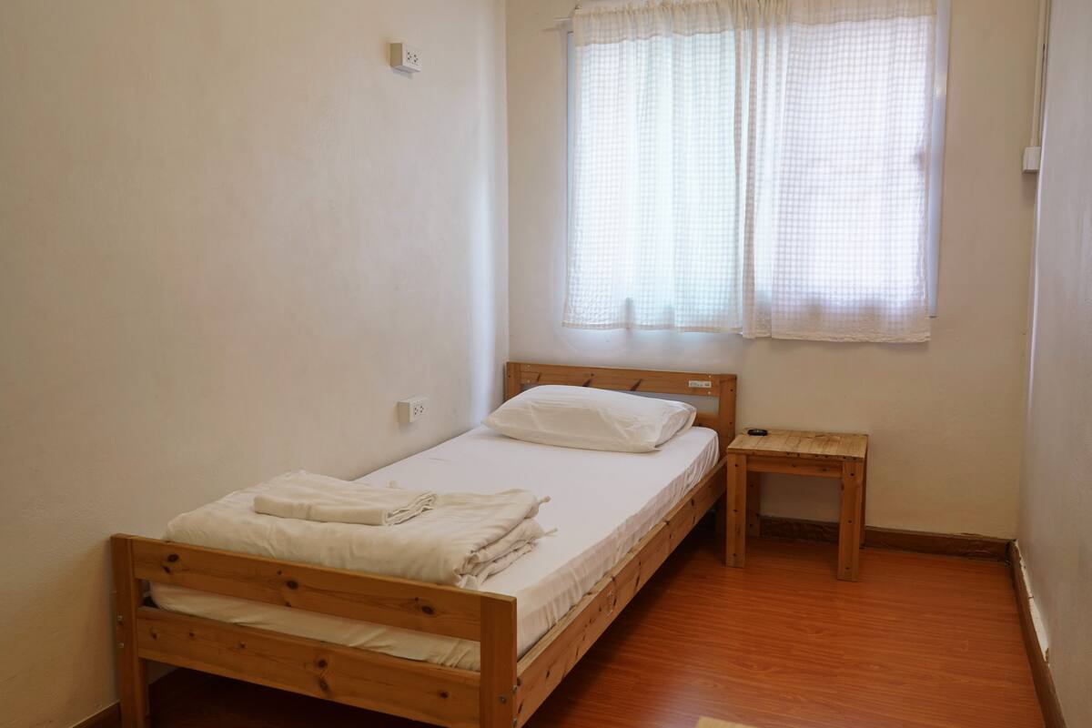 A single wooden bed with a white mattress and pillows, a small wooden nightstand, and a window with white curtains.