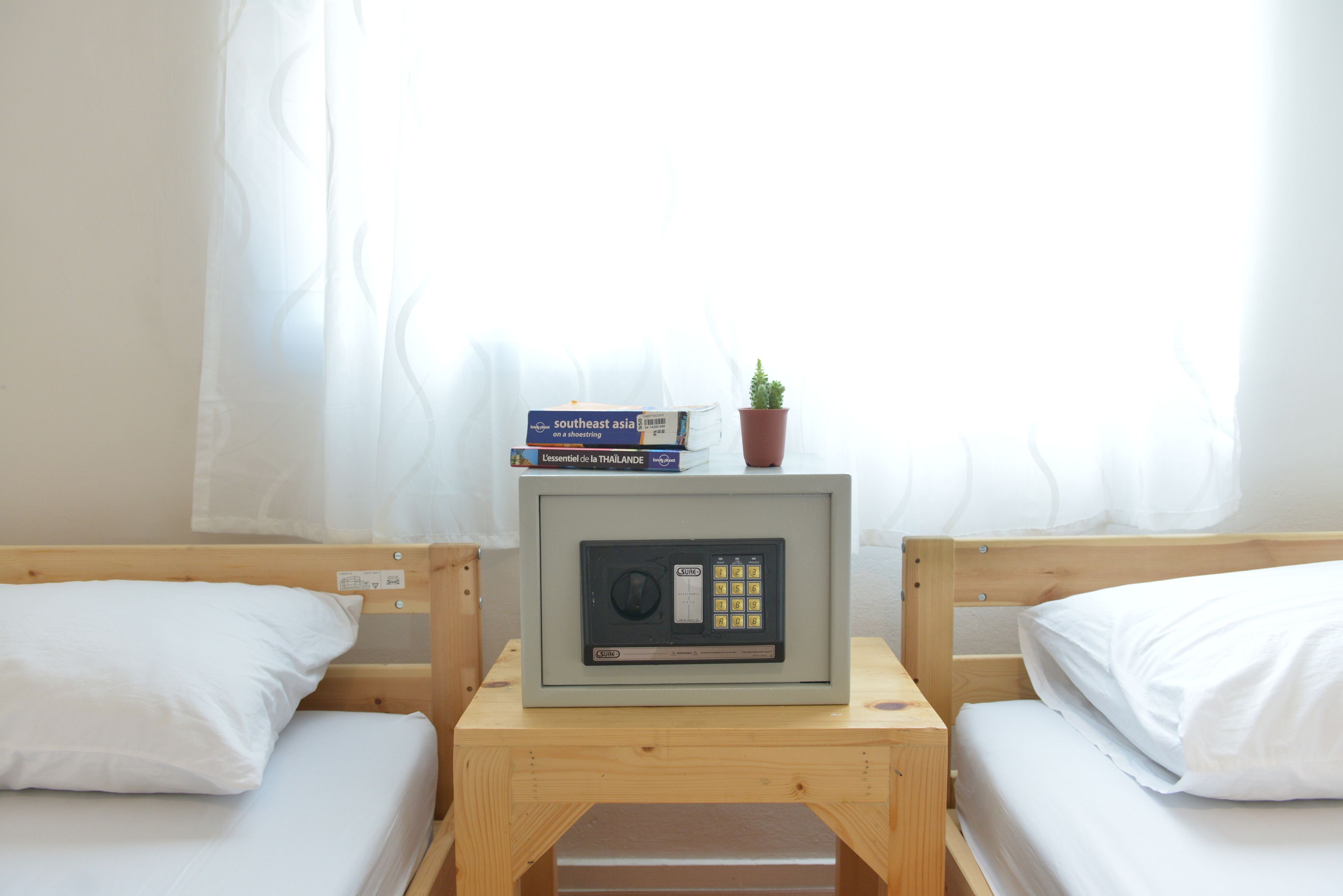 Una habitación pequeña con dos camas de madera, una mesita de noche con microondas y una ventana con cortinas traslúcidas.