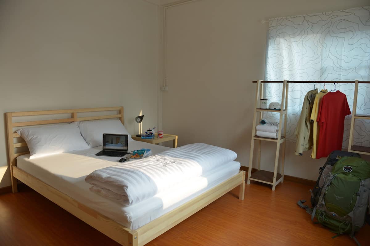 A single bed with a wooden frame, a bedside table with a lamp, a laptop, and a small shelf with items. There is a window with a view of a building and a clothing rack with folded clothes.