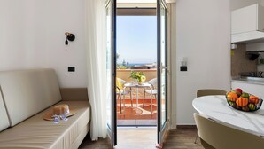A room with a sofa, a dining table with chairs, a kitchen area, and a view through an open door to an outdoor patio. - Villa Collina (Giardini Naxos)